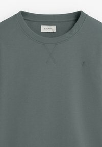 Felpa verde con scollo tondo a coste, tessuto strutturato e piccolo logo sul petto. Presenta un dettaglio a punto croce sullo scollo.