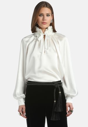 Blusa de satén blanca con cuello con volantes y cierre de lazo. Mangas largas con puños elásticos, combinada con pantalones de terciopelo negro y un cinturón trenzado.