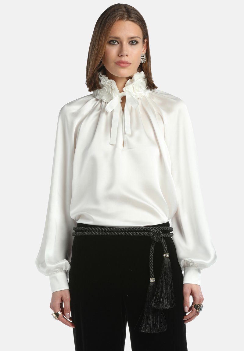 Blusa di raso bianco con scollatura a volant e chiusura a cravatta. Maniche lunghe con polsini elasticizzati, abbinata a pantaloni di velluto nero e una cintura intrecciata.