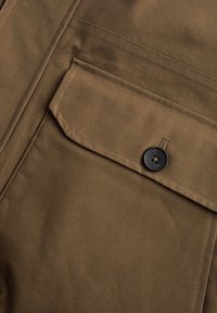 Rudas audinys kišenėje su uždengimu, kurį laiko vienas tamsus mygtukas, matomi detaliai siuvimo darbai.