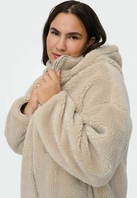 Bežs flīsu jaka ar kapuci, kam ir mīksta tekstūra, priekšpuses rāvējslēdzis un pārspīlēta piedurkne. Tai ir brīvā apgriezuma un ērta izskata.