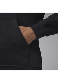 Mano con tono di pelle medio posizionata all'interno della tasca anteriore di un hoodie nero a maniche lunghe, su uno sfondo grigio.