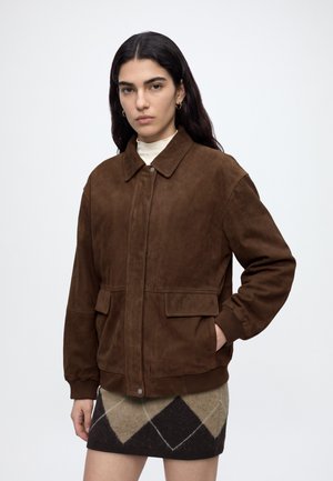 Woman with long dark hair wears brown suede jacket over white top and patterned brown and beige skirt, standing against plain background.