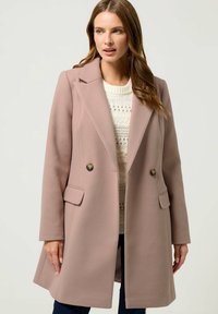 Dubbelzijdige jas van zacht roze stof, met grote revers, twee voorzakken en decoratieve gouden knopen. Getextureerde gebreide trui eronder.