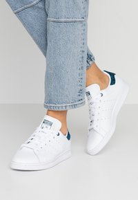 Zapatillas de cuero blancas con acentos perforados, lengüeta trasera con patrón verde y suela de goma blanca, combinadas con jeans de mezclilla azul claro.