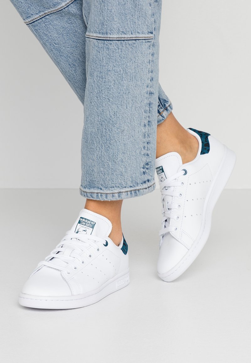 Zapatillas de cuero blancas con acentos perforados, lengüeta trasera con patrón verde y suela de goma blanca, combinadas con jeans de mezclilla azul claro.