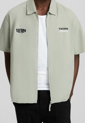 Mann trägt ein hellgraues kurzärmeliges Zip-Shirt über einem weißen Tanktop und schwarzen Hosen, mit "TAVERN Fuel Co." Logos auf der Brust.