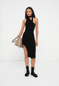 Μαύρο ριμπ midi φόρεμα με halter λαιμόκοψη και σχισμή στο πλάι. Το μοντέλο κρατά μια μπεζ τσάντα, φοράει μαύρες κάλτσες και επίπεδα παπούτσια.