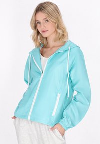 Giacca leggera con cappuccio di un luminoso color turchese, con chiusura a zip e tasche, dotata di polsini elasticizzati e una texture liscia e lucida.