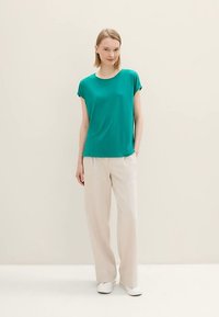 Maglietta a maniche corte turchese abbinata a pantaloni beige a gamba larga e scarpe da ginnastica bianche, su uno sfondo neutro. Design minimalista.