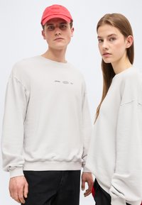 Νεαρός άνδρας με κόκκινο καπελάκι και λευκό φούτερ στέκεται δίπλα σε νεαρή γυναίκα με μακριά μαλλιά που φοράει λευκό μεγάλο φούτερ, και οι δύο κοιτούν μπροστά.