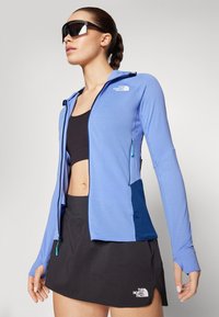 Jacheta sport cu fermoar de culoare bleu deschis, cu un finisaj texturat, purtată împreună cu un top scurt negru și o fustă sport neagră, având un logo pe fustă.