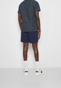 Mężczyzna stoi tyłem, ubrany w ciemną koszulę z krótkim rękawem w wzory, granatowe szorty, białe skarpety do kostek i białe sneakersy z czarnymi akcentami.