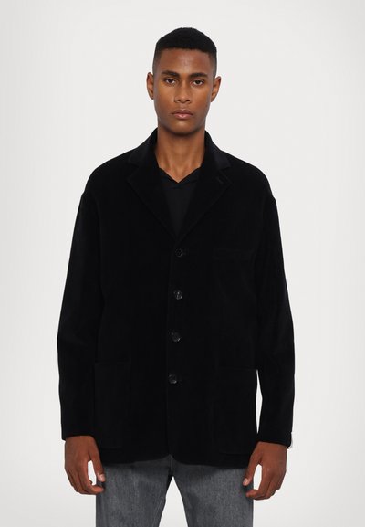 Black corduroy jacket with a notched collar, front button closure, and two large patch pockets. Features a relaxed fit and textured fabric.