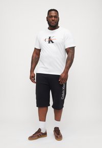 Hvit bomull T-skjorte med "Calvin Klein Jeans" logo i svart og oransje, kombinert med svarte bomullsshorts med logo på siden, brune sneakers.