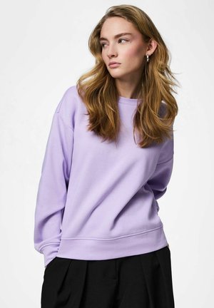 Jeune femme aux cheveux blonds ondulés portant un sweat-shirt lavande et un pantalon noir, regardant sur le côté sur un fond uni.