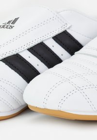 Vita sportskor med svarta ränder och vadderad yta. Har en slät yta och en gummisula. Logotyp synlig på sidan.