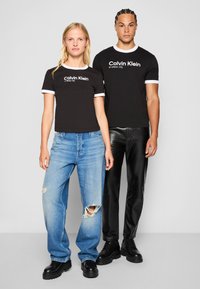 Juodos medvilnės marškinėliai su baltais apvadais ir užrašu "Calvin Klein endless city" priekyje, dėvimi su suplėšytais mėlynais džinsais ir juodomis kelnėmis.