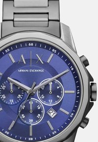 Roostevaba terasest kell sinise taustaga, millel on hõbedased tunni markerid, kolm alalise ajaga näidikut ja kuupäevaaken. Kaheksanurkne korpus ja linkidega randmepael.