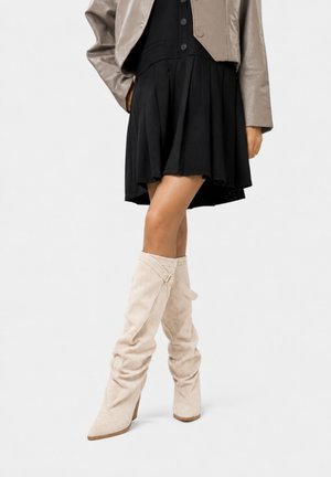 Kvinde iført sort plisseret nederdel, taupe cropped jakke og lyse beige løstsiddende knæhøje støvler med lille spædedetalje.