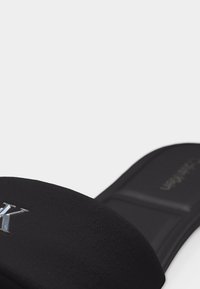 Μαύρη παντόφλα με φαρδιά υφασμάτινη λωρίδα που φέρει ασημένιο λογότυπο "K" σε λευκό φόντο.