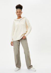 Crème kabelgebreide trui met een halve ritssluiting, gecombineerd met lichtkhaki wijde broekspijpen en witte sneakers, wat een relaxte pasvorm laat zien.