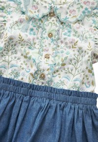 Tenue enfant avec blouse à motif floral présentant des bords festonnés et des boutons en bois, associée à une jupe en denim bleu froncée.
