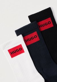 Kolme paria sukkia väreissä valkoinen, laivastonsininen ja musta. Sukissa on resorikuvio ja punainen suorakaiteen muotoinen etiketti, jossa lukee "HUGO" mustilla kirjaimilla.