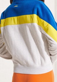 Bluziak polarowy w blokach kolorystycznych, w odcieniach niebieskiego, żółtego i szarego. Posiada wysoki kołnierz, ściągacz u dołu oraz miękką teksturę.