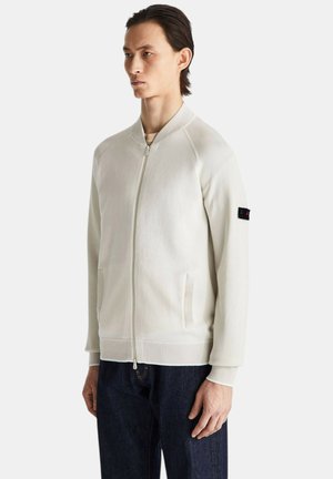 Mężczyzna w białej bomberce na suwak z prążkowanymi mankietami i kołnierzem, stojący na jednolitym tle.