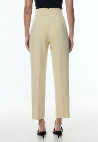 Beige high-waisted broek met een getailleerde pasvorm, met een gladde textuur en een subtiele achternaad, gecombineerd met zwarte sandalen met banden.
