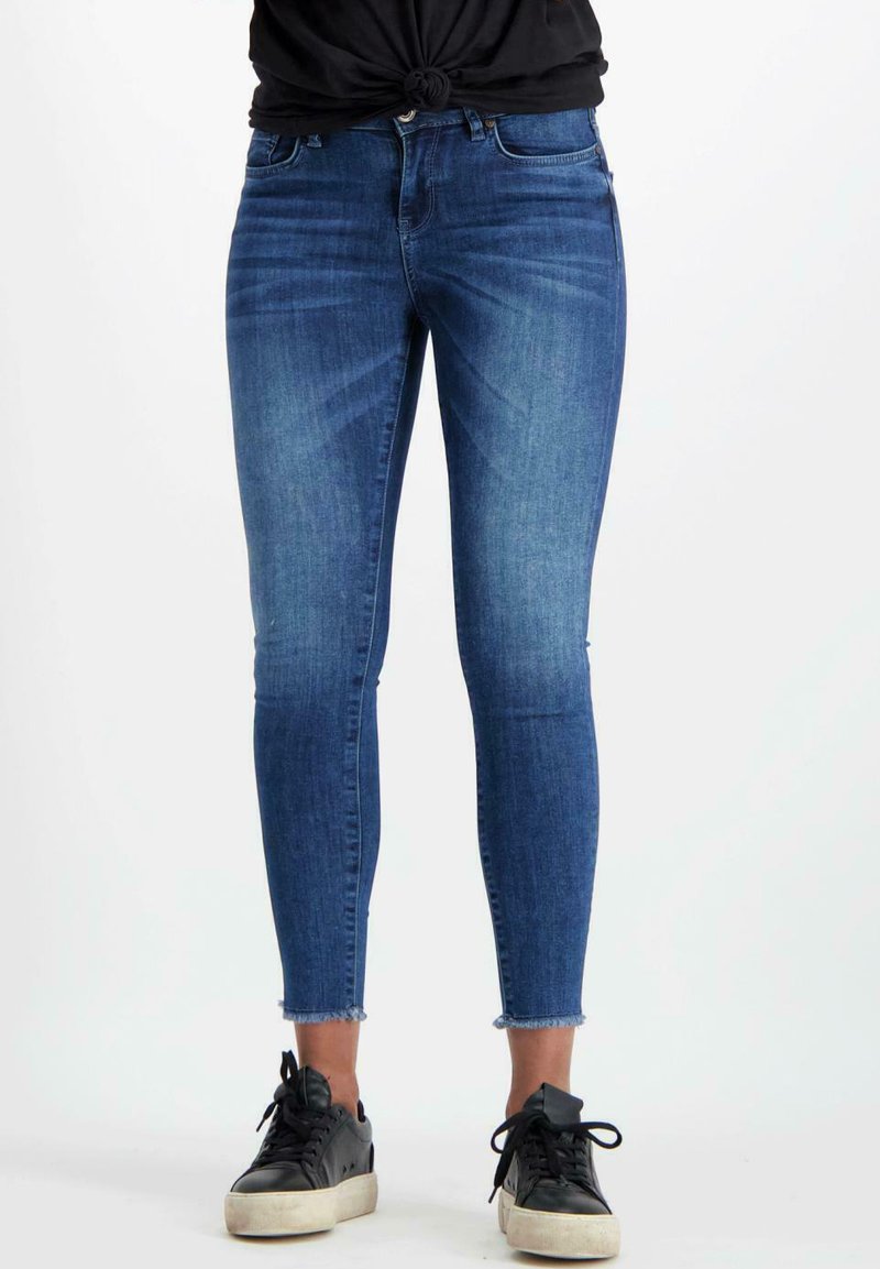 Dunne blauwe denim jeans met een rafelige zoom en een middenhoogte taille, gecombineerd met zwarte sneakers, met een gladde textuur en minimale details.