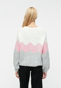 Adīta džemperis baltā, rozā un pelēkā krāsā ar zickzack rakstu; tam ir garas piedurknes un brīvs piegriezums. Mīksta tekstūra.