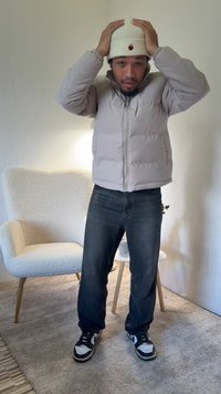 Világosszürke steppelt dzseki elülső cipzárral és oldalzsebekkel, sötét farmernadrággal és fekete-fehér tornacipővel párosítva. Fehér bordázott sapka.