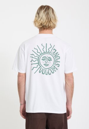 Blond persoon die een wit T-shirt draagt met een groen zonontwerp en de tekst "Volcom Stone" op de achterkant, gecombineerd met donkerbruine broek.