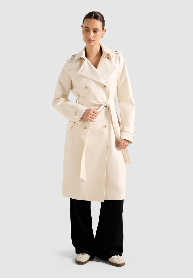 Donna che allaccia la cintura di un trench coat beige, indossando pantaloni neri a gamba larga e mocassini crema, in piedi su uno sfondo grigio chiaro.