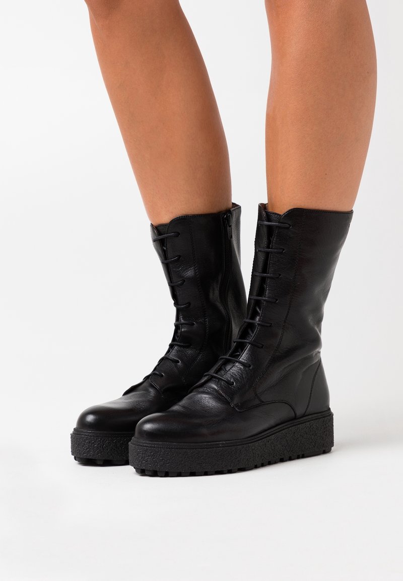 Jambes portant des bottes à plateforme en cuir noir mi-mollet avec des fermetures éclair sur les côtés, des orteils ronds et des semelles en caoutchouc texturé sur un fond blanc.