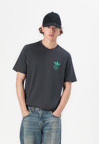 T-shirt en coton gris à manches courtes avec un motif graphique vert ; porté avec un jean en denim bleu et une casquette de baseball noire.