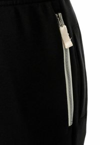Zwarte sweatpants met een zijzak met een crèmekleurige rits. De stof is zacht en textuur, wat zorgt voor een casual uitstraling.