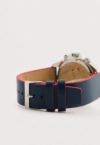 Cinturino in pelle blu navy con bordi rossi e fibbia argentata, attaccato a un orologio cronografo argentato posato su una superficie bianca.