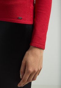 Maglione fitted rosso con maniche lunghe, texture a coste e un piccolo logo metallico all'orlo, abbinato a un capo nero.