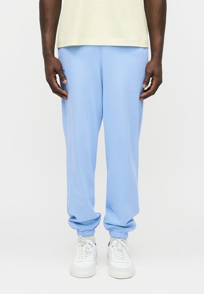 Egy férfi világoskék, elasztikus szegélyű jogger nadrágot, fehér tornacipőt és halványsárga rövid ujjú inget visel, fehér háttér előtt áll.