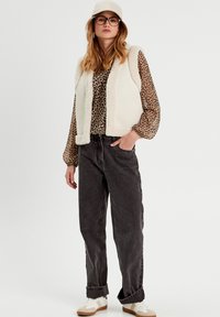 Chaleco de piel sintética en crema, camisa con estampado, jeans negros de cintura alta y zapatillas blancas con detalles en marrón; conjunto informal con texturas superpuestas.