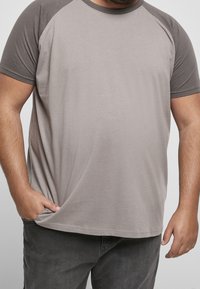 Man i grå raglanärmad t-shirt med ena handen i fickan och den andra vilande vid sin sida mot en enkel bakgrund.