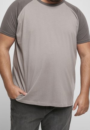 Man i grå raglanärmad t-shirt med ena handen i fickan och den andra vilande vid sin sida mot en enkel bakgrund.