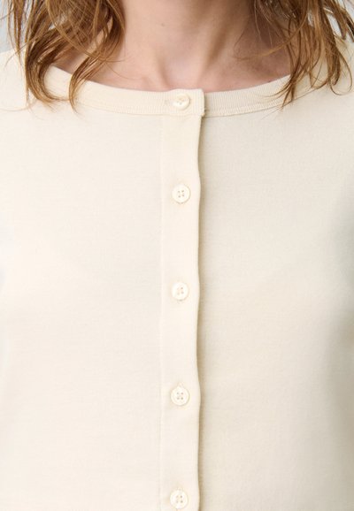 Femme portant un cardigan boutonné de couleur crème avec un col rond, mettant en évidence sa clavicule et des mèches de cheveux.