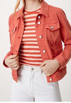 Kobieta w koralowej kurtce dżinsowej na czerwonej i beżowej pasiastej koszuli, w połączeniu z białymi, wysokim stanem jeansami, poprawiająca kurtkę.