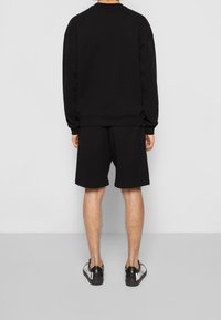 Osoba stojící zády k fotoaparátu, oblečená v černém dlouhém rukávu, černých šortkách a černých teniskách s bílým nápisem na jednobarevném pozadí.