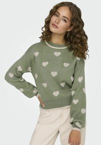 Žalias megztinis su grietinėlės spalvos širdelėmis. Turi apvalią iškirptę, nusileidusius pečius ir elastines rankoves bei apačią.