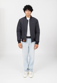 Zwarte bomberjack met geribbelde manchetten en ritszakdetails, gecombineerd met lichtblauwe jeans en witte sneakers op een effen achtergrond.